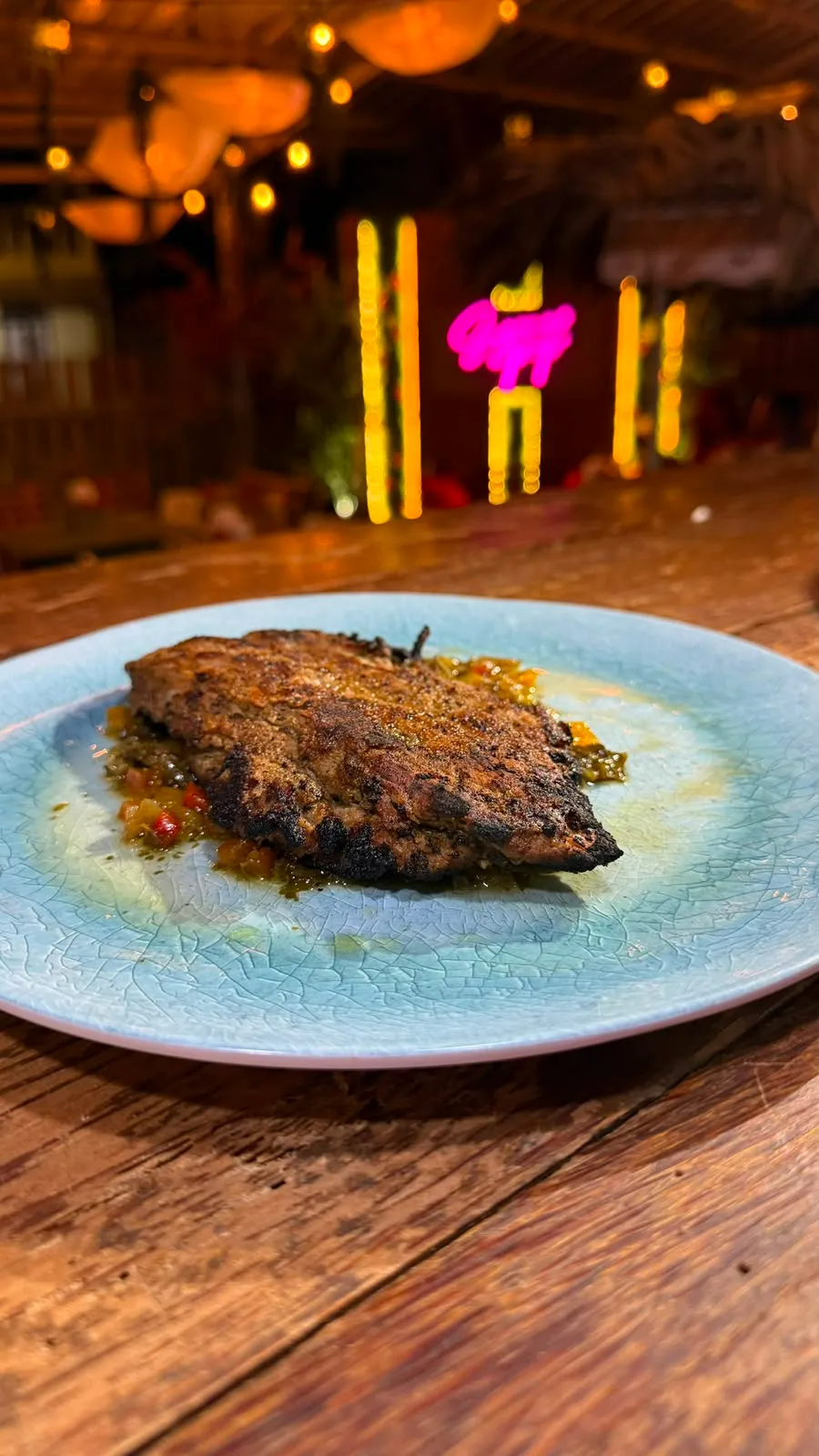 Churrasco a la parrilla sobre vegetales, con el neón de Sazonao Pop al fondo.